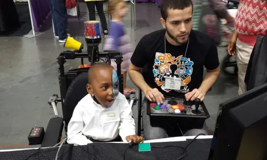A photo of a child in a power-chair reacting positively to a video game being played by a man using an assistive controller. Image via AbleGamer's official site.